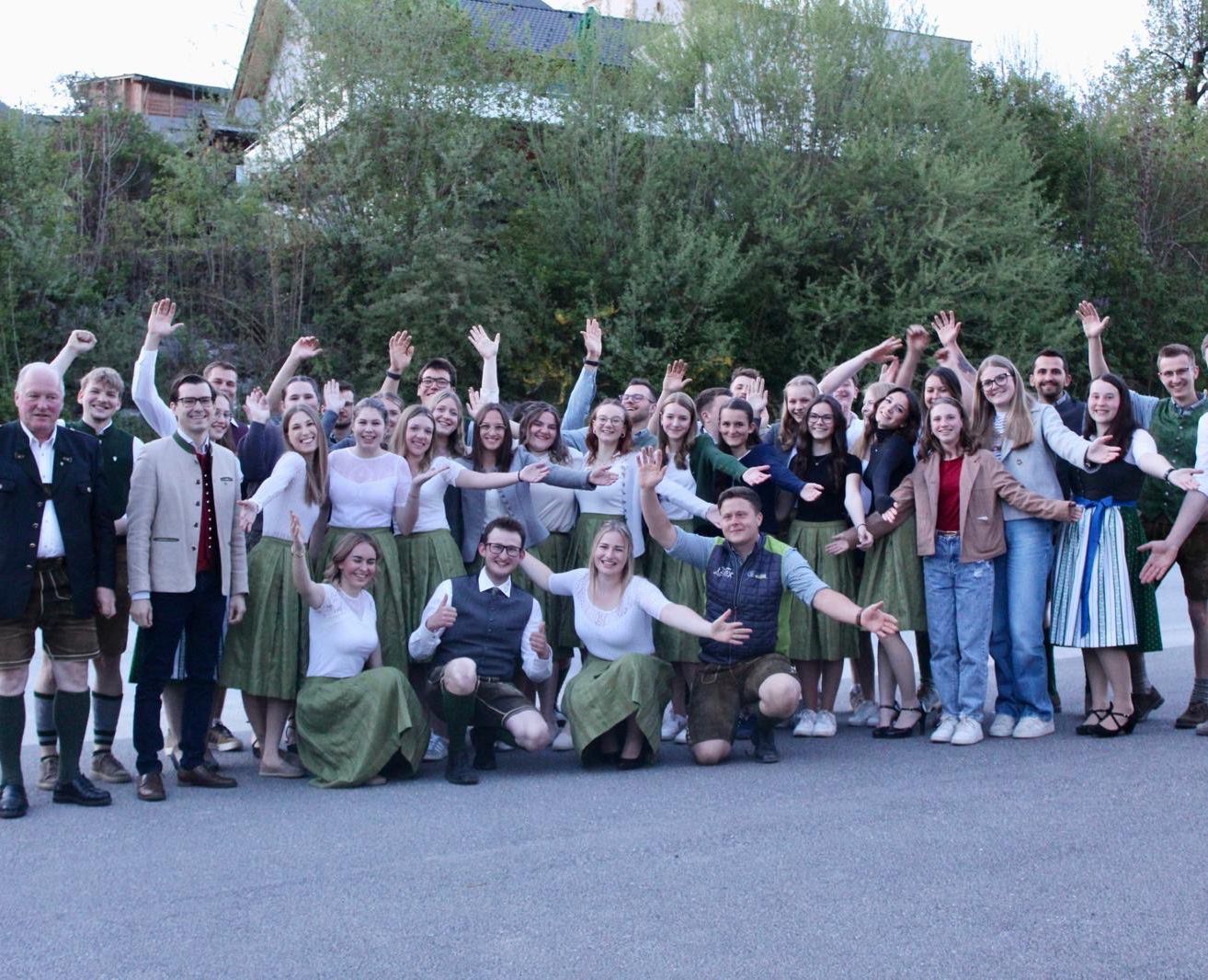 Eine große Gruppe von Menschen posiert für ein Foto, einige tragen traditionelle Kleidung. Sie stehen auf einer Straße mit Bäumen im Hintergrund.