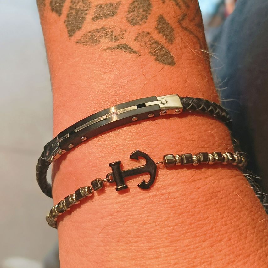 Ein Nahaufnahme des Handgelenks zeigt zwei Armbänder, eines mit Silberverschluss und schwarzem Leder und eines mit einem Anker und Perlen.