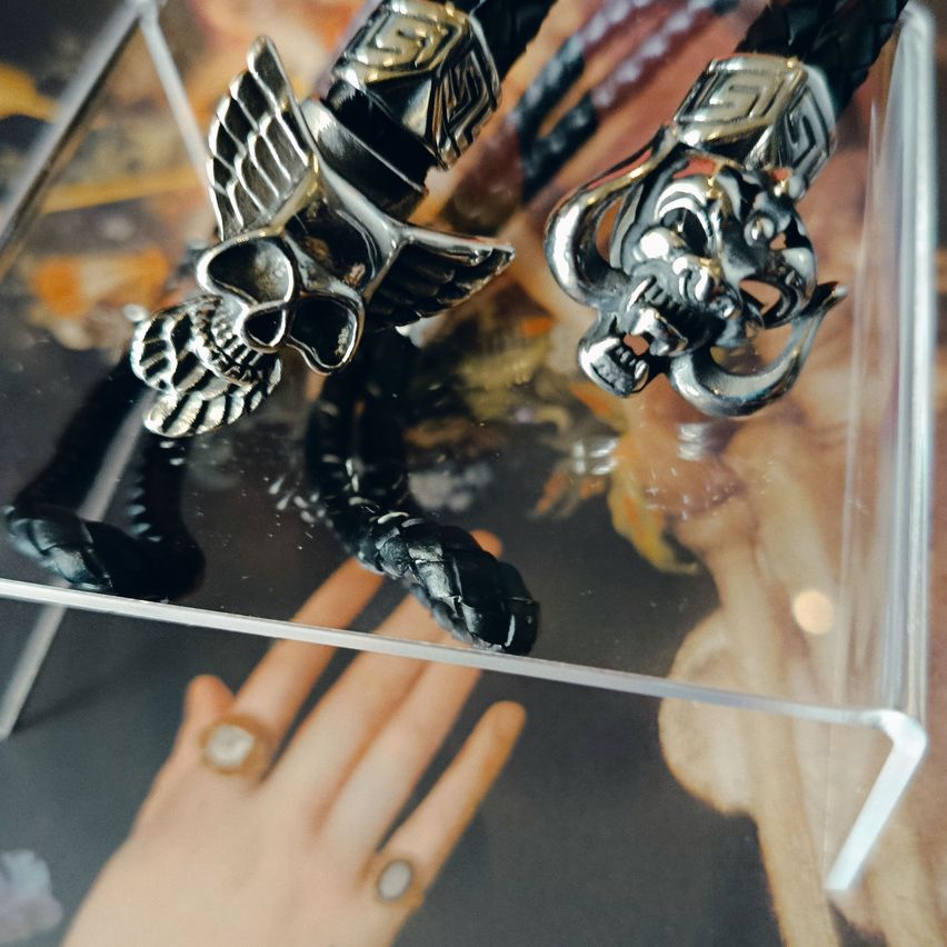 Zwei schwarze Lederarmbänder mit Metallschädeln, Flügeln und einer Schlange in einer Glasschatulle. Unten ist eine Hand mit Ringen sichtbar.