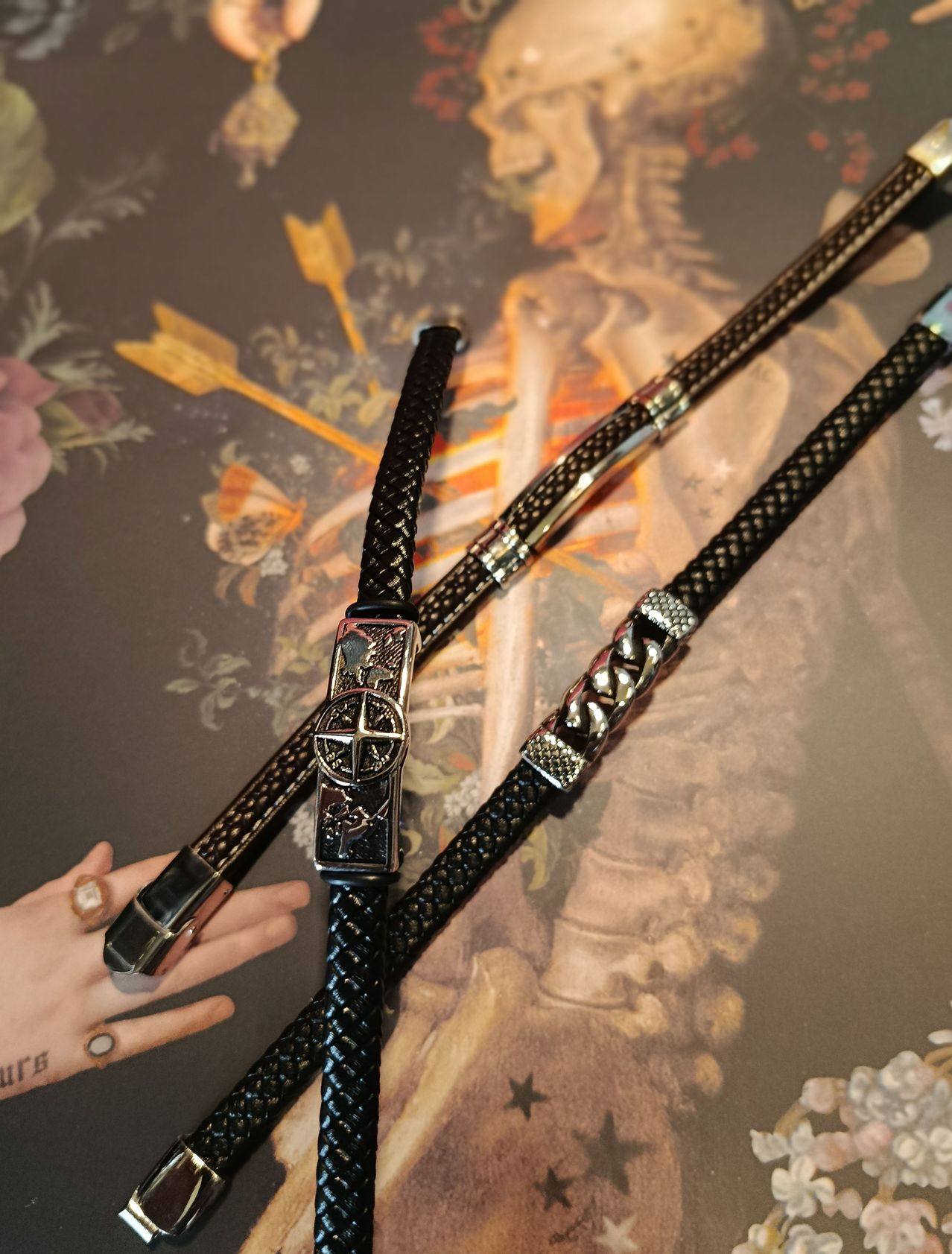 Zwei schwarze Lederarmbänder mit silbernen Schnallen liegen auf einer strukturierten Oberfläche. Ein Armband hat ein kompassähnliches Design.