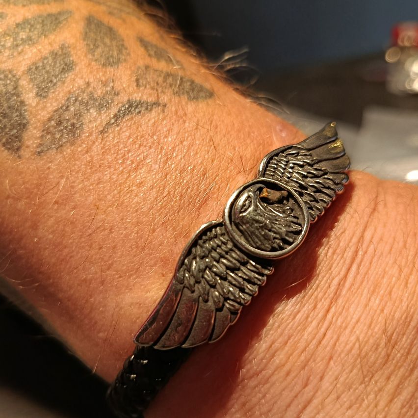 Ein Armband mit einem silbernen Adlerdesign wird am Arm einer Person mit einer Tätowierung getragen. Das Armband wird nah an der Haut getragen.