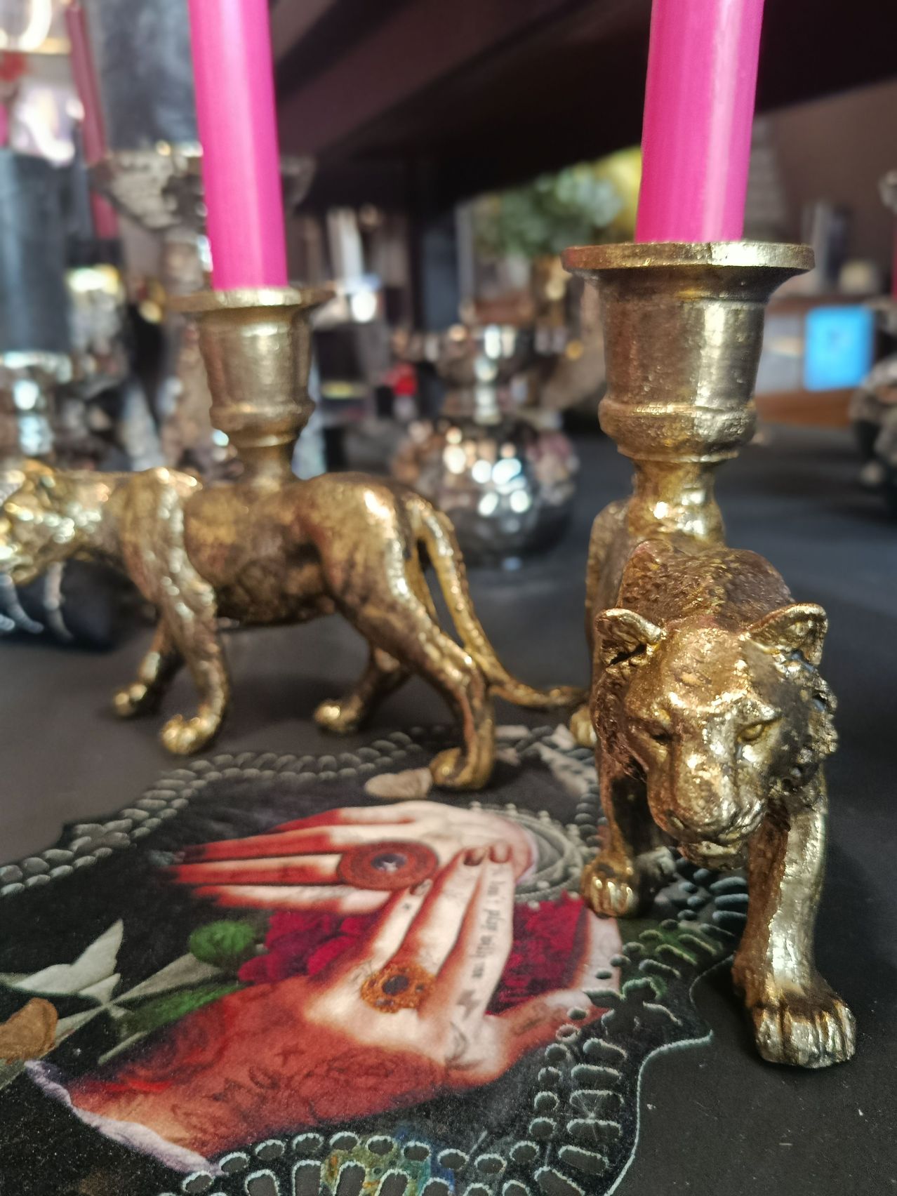 Zwei goldene Löwenfiguren halten rosa Kerzen auf einem Tisch, umgeben von einem mystischen Kunstwerk. Im Hintergrund fügen verschiedene metallische Objekte zur mystischen Atmosphäre bei.