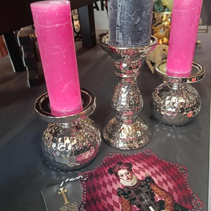 Drei Kerzenständer mit silbernen Basen, zwei mit rosa Kerzen und einer mit einer schwarzen Kerze. Ein dekoratives Tuch mit dem Porträt einer Frau im Vordergrund.