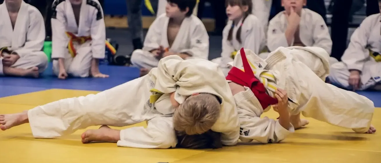 Zwei Kinder in weißen Uniformen sind in einem Judokampf verwickelt, wobei eines oben liegt. Zuschauer, darunter Kinder, schauen von den Seitenlinien zu. Der Boden ist gelb.