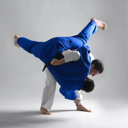 Zwei Männer in blauen und weißen Kampfsportuniformen führen eine dynamische Judo-Bewegung vor einem weißen Hintergrund aus. Ein Mann wird gehoben und der andere legt eine Beinhaltung an.