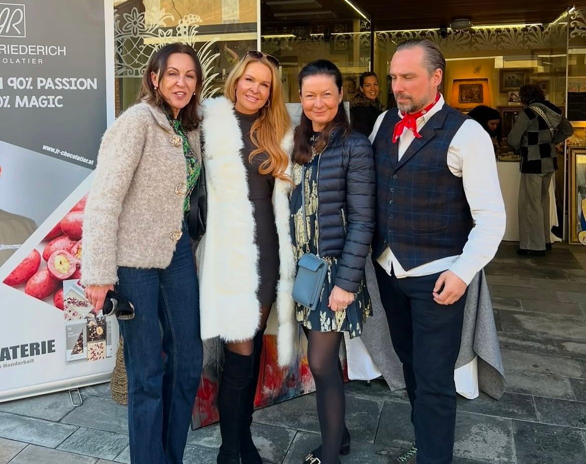 Vier Personen stehen vor einem Geschäft und lächeln für ein Foto. Ein Mann trägt ein rotes Halstuch und eine Weste. Hinter ihnen spiegelt eine Glastür Menschen und Gegenstände.