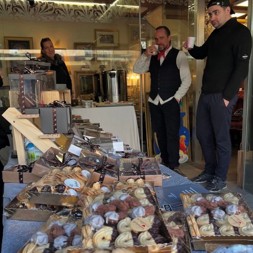 Zwei Männer stehen in einem Geschäft, einer trinkt aus einer Tasse und der andere hält eine Tasse. Hinter ihnen steht ein Tisch mit vielen Geschenkboxen. Eine weitere Frau steht in der Nähe des Tisches.