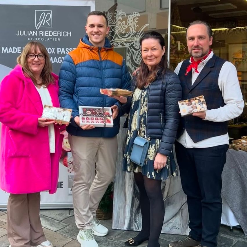 Vier Personen posieren für ein Foto vor einem Schild von Julian Riederich Chocolatier, jeder hält eine Schachtel Pralinen. Sie lächeln und scheinen ein Ereignis zu feiern.