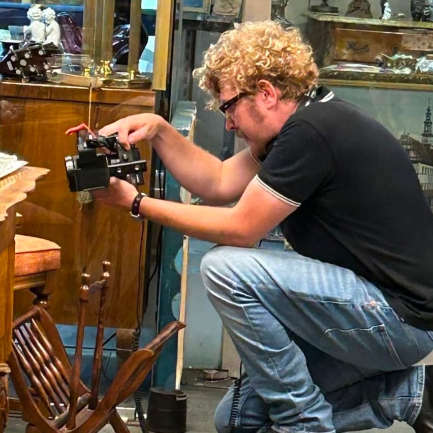 Ein Mann mit lockigen Haaren kniet und hält ein Gerät in den Händen. Er trägt eine Brille und Kopfhörer. Hinter ihm steht ein hölzerner Schrank mit einigen Gegenständen darauf.
