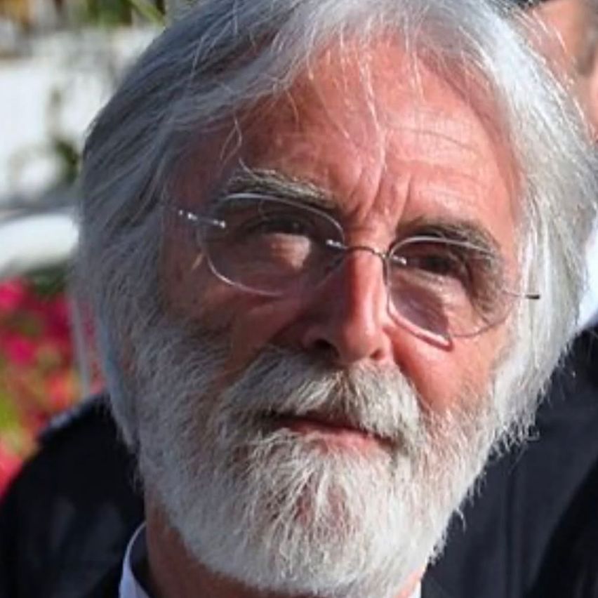 Ein älterer Mann mit weißem Haar und Bart, der eine Brille trägt, scheint auf etwas fokussiert zu sein. Der Hintergrund ist unscharf.