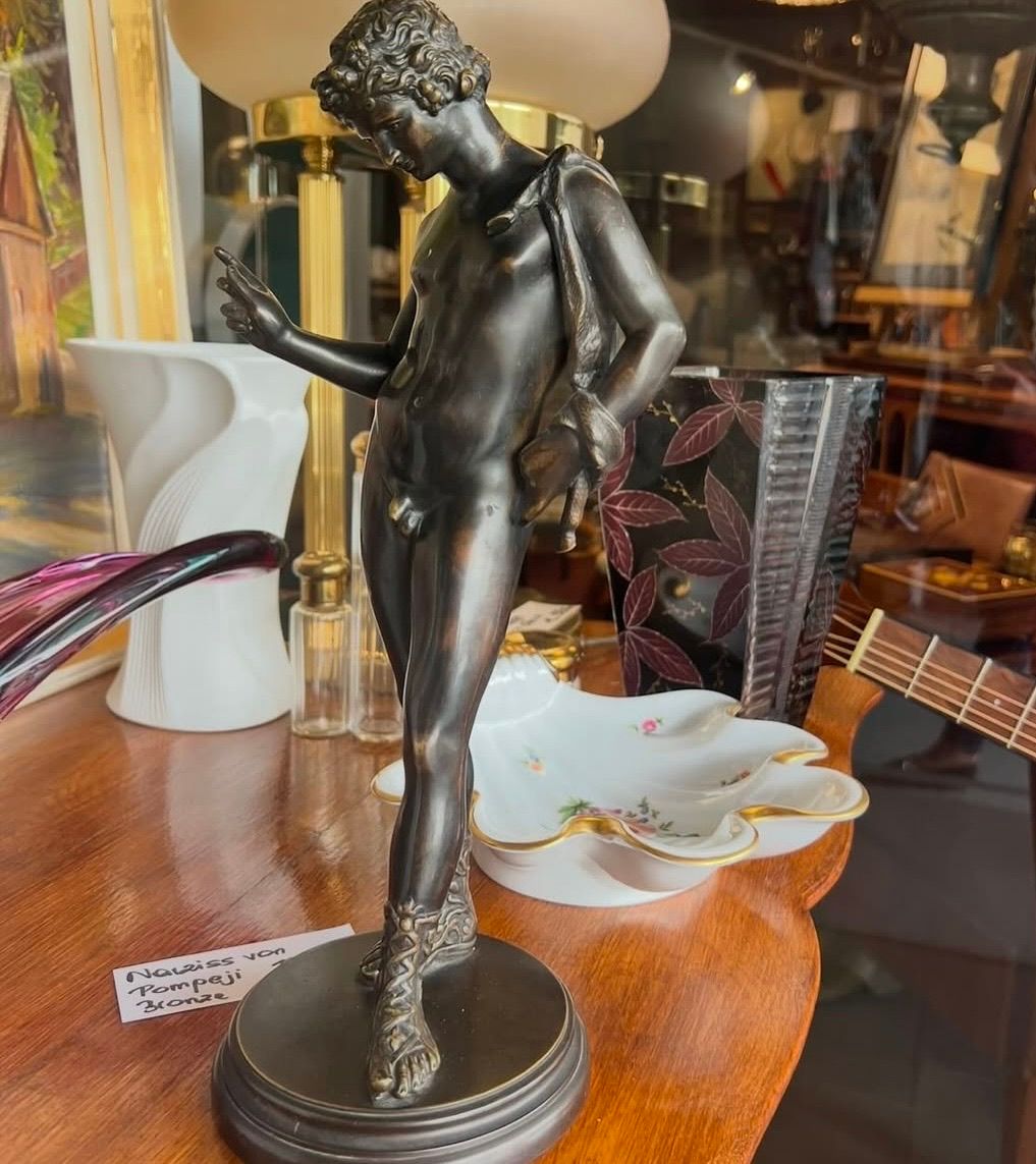 Eine Bronzestatue steht auf einem Holztisch mit einer Keramikschale, einer Gitarre und anderen Gegenständen. Die Statue zeigt eine männliche Figur mit Sandalen und einer Umhängetasche.
