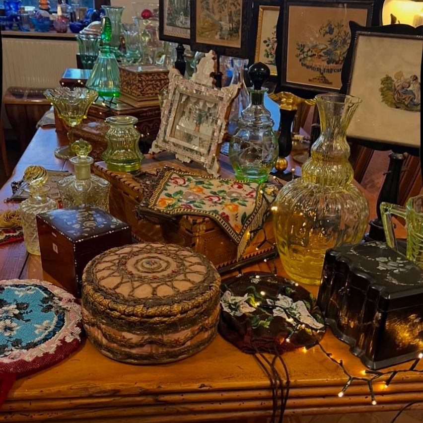 A wooden table displays various decorative items: vases, boxes, picture frames, and a lamp. The table is illuminated by fairy lights. The background shows a radiator and additional framed pictures.