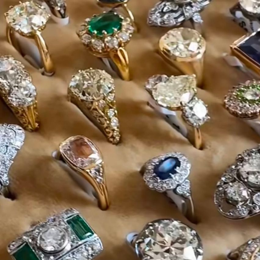 A collection of various rings with different gemstones on a brown surface. Each ring is uniquely designed, featuring diamonds, emeralds, and sapphires. The arrangement showcases different sizes and styles.