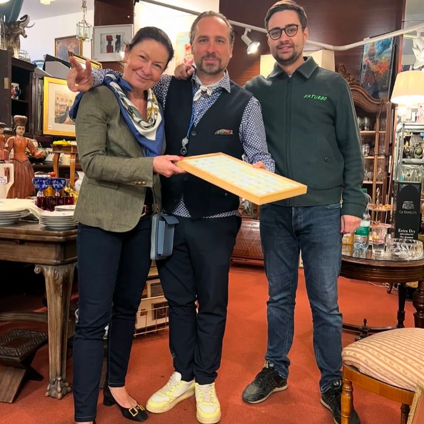 Three people stand in a room, smiling. A woman and a man holding a wooden tray, and another man beside them. Behind them are shelves with various items. A desk has plates and a teapot. A lamp is on the right.