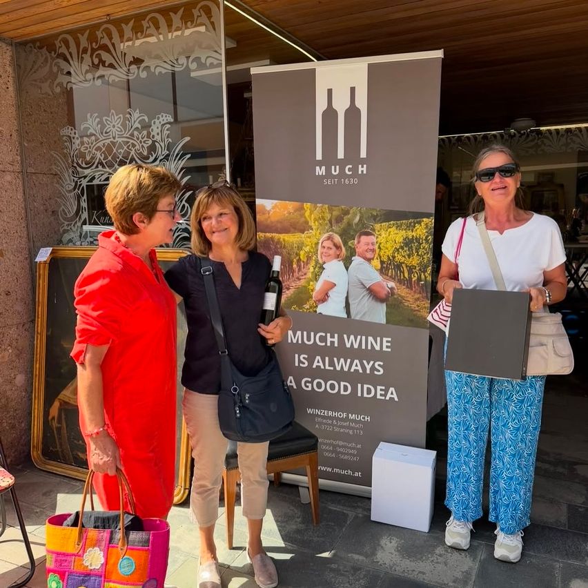 Three women stand in front of a wine advertisement banner, smiling and holding a bottle of wine and a box. The banner features a vineyard and the slogan 'Much wine is always a good idea.'