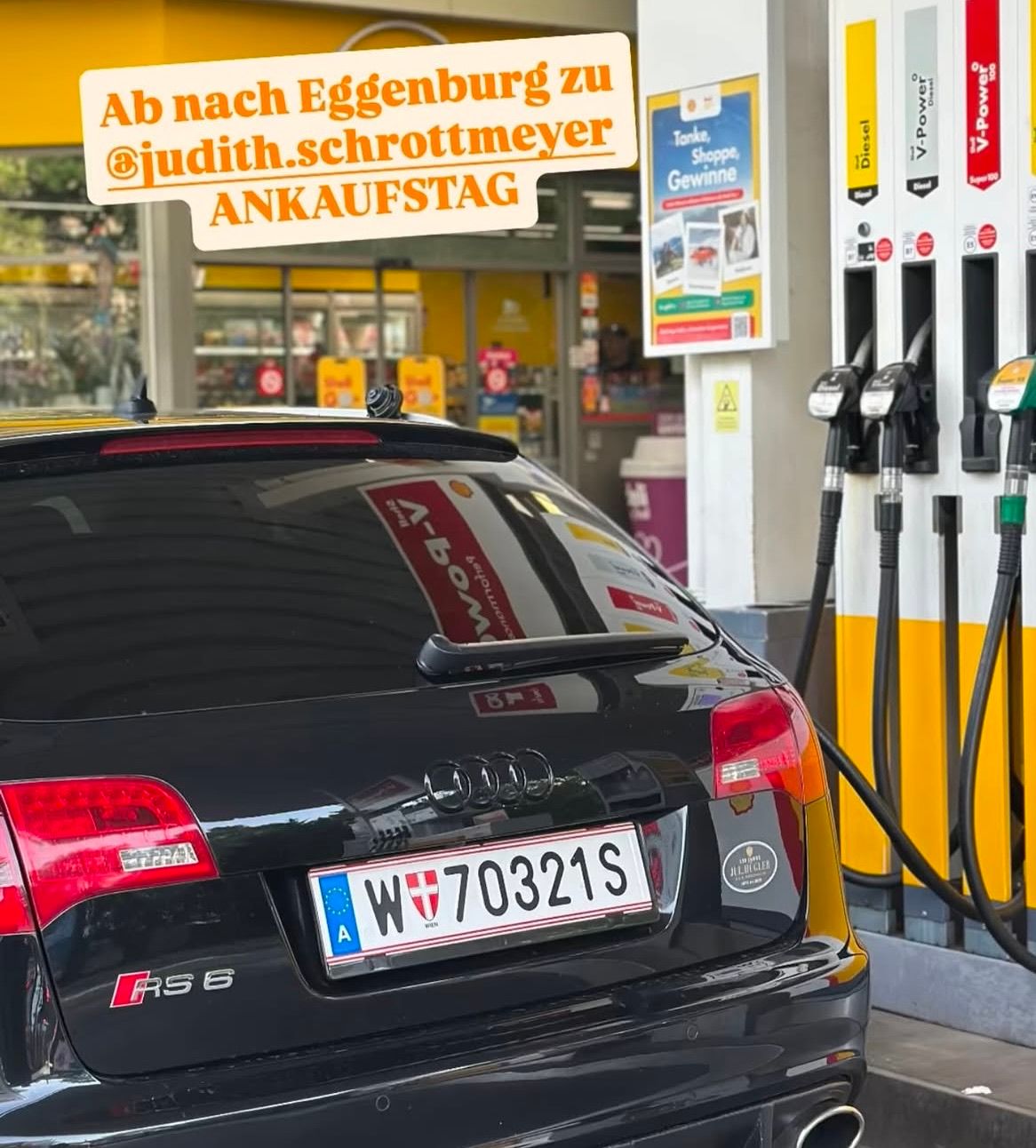 A black Audi RS6 is parked at a gas station with a fuel pump attached. The car has a license plate with the number W 70321S. Above the car, there is a signboard with the text 'Ab nach Eggenburg zu @judith.schrottmeyer ANKAUFSTAG'.