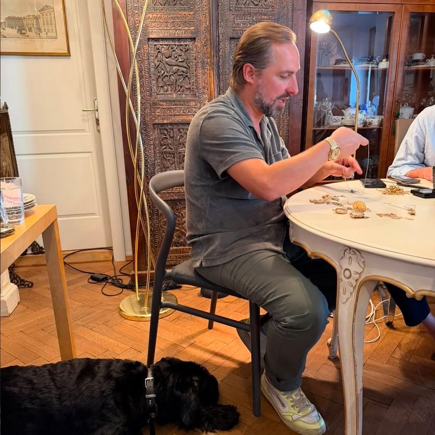 A man is sitting at a table with various items on it, wearing a gray shirt and pants. He is holding a small object and looking at it. A dog is lying on the floor next to him.