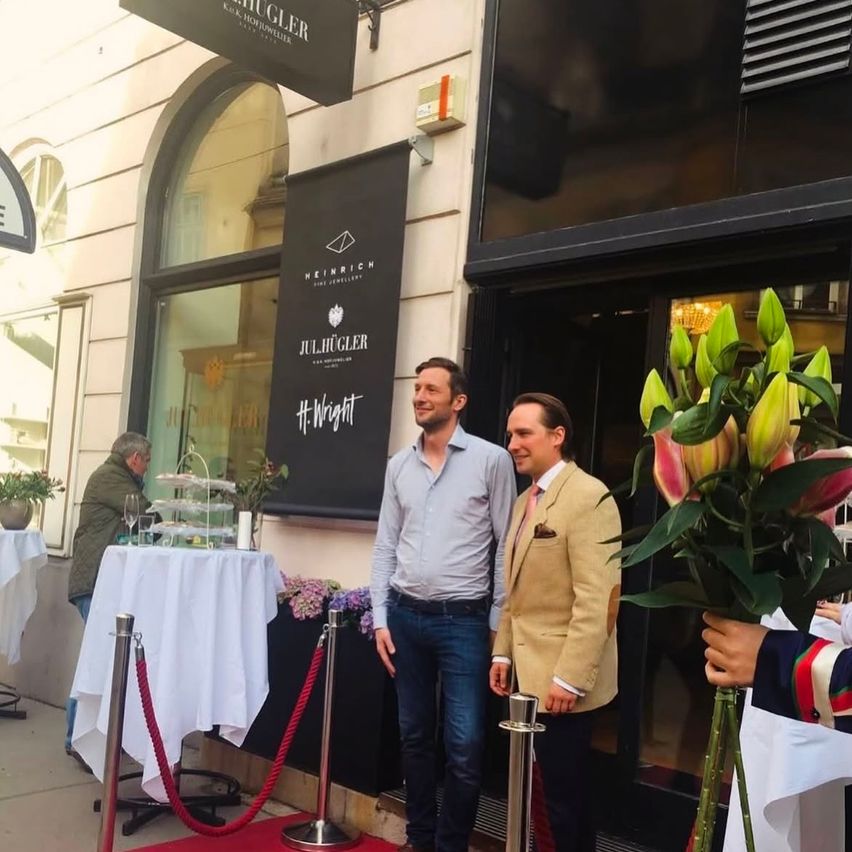 Two men stand outside a store, smiling, with flowers in front of them. A table with a white tablecloth and a person in the background.
