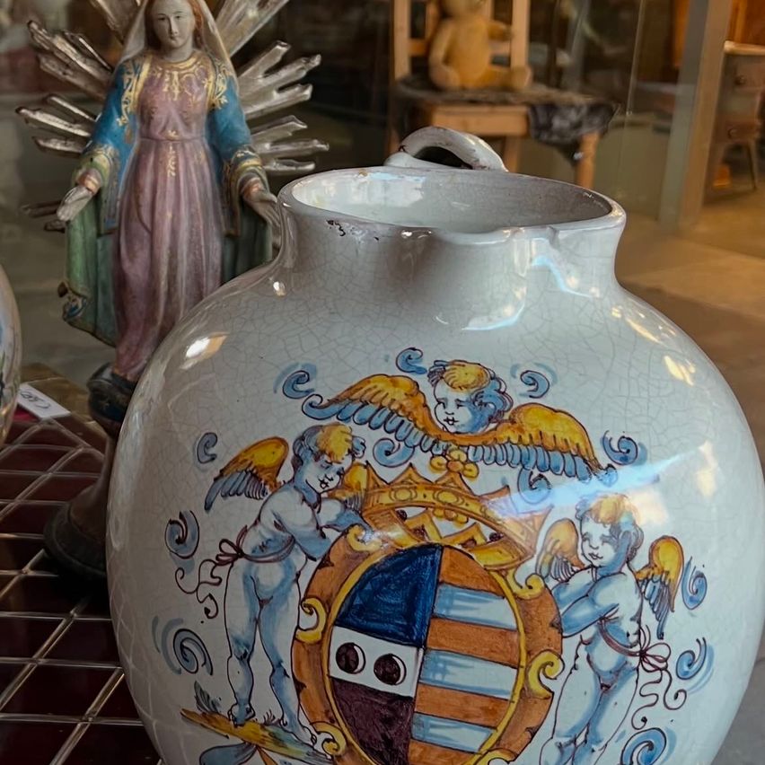 A decorative ceramic vase with an emblem of two cherubs holding a crown over a shield sits on a tiled surface. Behind it, a statue of the Virgin Mary is displayed in a glass case.