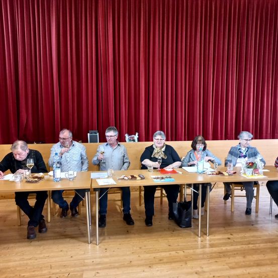 Eine Gruppe älterer Menschen sitzt an einem langen Tisch in einem Raum, hält Weingläser und einige von ihnen trinken.