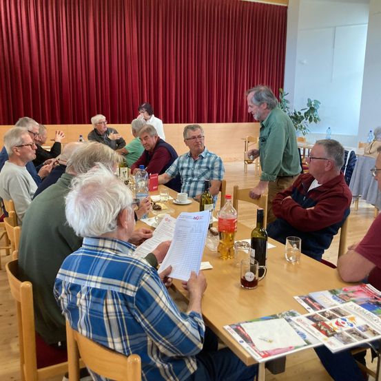 Mehrere ältere Männer sitzen um einen Holztisch in einem Raum, möglicherweise während einer Besprechung. Ein Mann liest ein Dokument. Flaschen, Becher und Papiere sind auf dem Tisch. Ein anderer Mann steht hinter ihnen, und eine Gardine ist im Hintergrund.