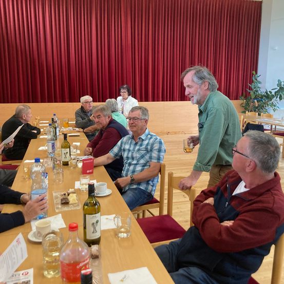 Eine Gruppe von Menschen sitzt um einen langen Tisch herum, lächelt und unterhält sich. Flaschen, Tassen und Teller stehen auf dem Tisch. Ein Mann in einem grünen Shirt steht vor ihnen.