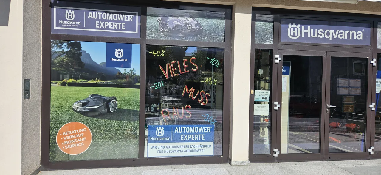 Ein Schaufenster zeigt einen Rasenmäher mit verschiedenen Rabatten in Rot auf der Scheibe. Das Schild sagt 'AUTOMOWER EXPERTE' und beinhaltet ein Husqvarna-Logo.