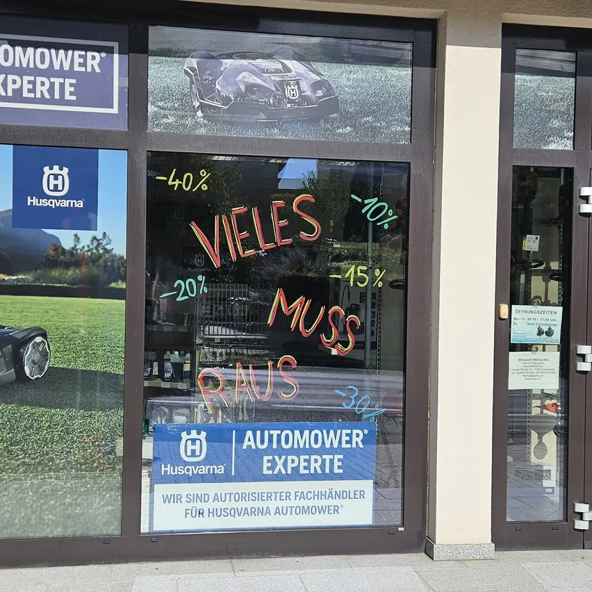 Ein Schaufenster zeigt einen Rasenmäher mit verschiedenen Rabatten in Rot auf der Scheibe. Das Schild sagt 'AUTOMOWER EXPERTE' und beinhaltet ein Husqvarna-Logo.