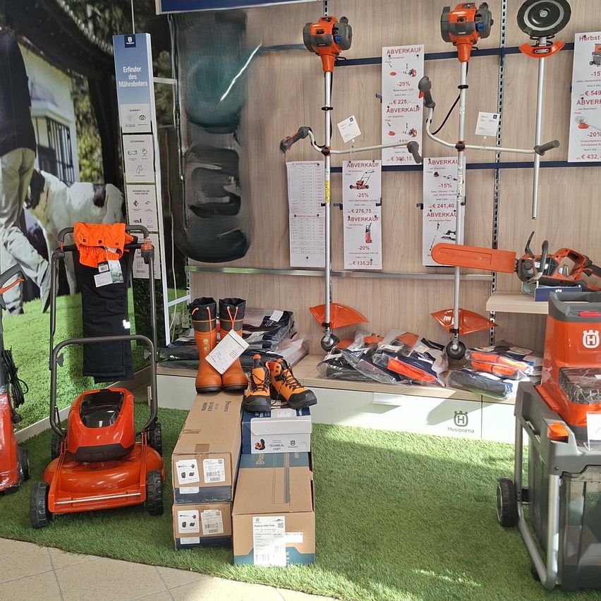 Eine Ausstellung von Husqvarna-Gartengeräten in einem Geschäft, einschließlich Rasenmäher, Trimmer und Schutzausrüstung. Die Produkte sind auf Regalen und am Boden angeordnet.