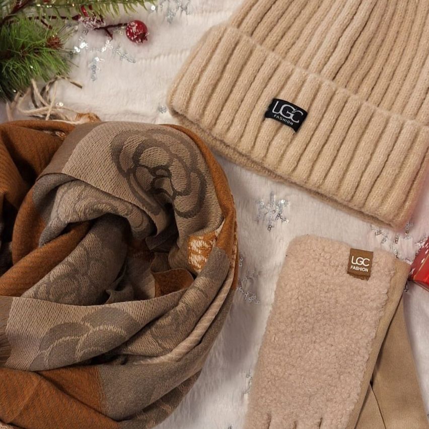 Ein beige Strickmütze, Schal und Handschuhe sind auf einer weißen Oberfläche ausgestellt. Die Mütze hat ein Label mit der Aufschrift 'LGC FASHION'. Der Schal hat ein Muster. Die Handschuhe sind gefaltet.
