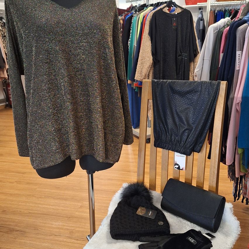 Ein Mannequin präsentiert ein metallisches Oberteil in einem Bekleidungsgeschäft mit verschiedenen Kleidungsstücken an Kleiderbügeln im Hintergrund. Auf einem Holzstuhl liegen eine schwarze Handtasche, eine Mütze und Handschuhe.