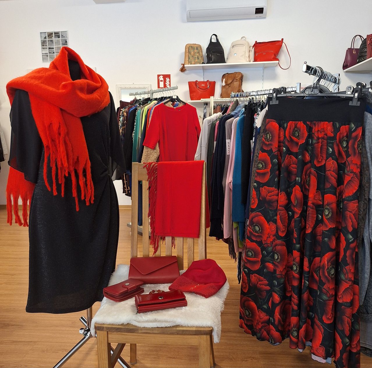 Ein Bekleidungsgeschäft zeigt ein schwarzes Kleid mit einem orangefarbenen Schal, einem roten Hut und roten Handtaschen. Ein roter Blumenrock und verschiedene Handtaschen hängen an Kleiderbügeln.