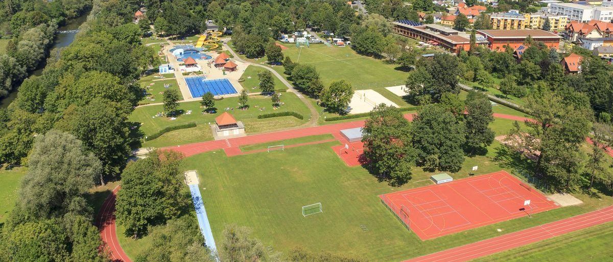 Luftaufnahme eines Sportkomplexes mit Schwimmbad, Fußballfeld, Laufbahn und Tennisplätzen.