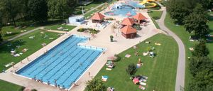 Luftaufnahme eines großen Freibadbereichs mit mehreren Schwimmbecken, darunter ein großes Schwimmbecken und kleinere. Es gibt Menschen, die schwimmen, sonnenbaden und sich auf dem Gras entspannen. Es gibt auch mehrere kleine Hütten und eine gelbe Wasserrutsche.
