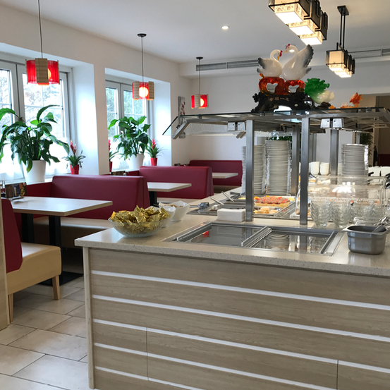 Ein Restaurant mit moderner Einrichtung, mit einem Buffet-Counter mit Tellern, Schüsseln und dekorativen Schwänen. Es gibt Tische mit Stühlen, Topfpflanzen und Lampen, die von der Decke hängen.