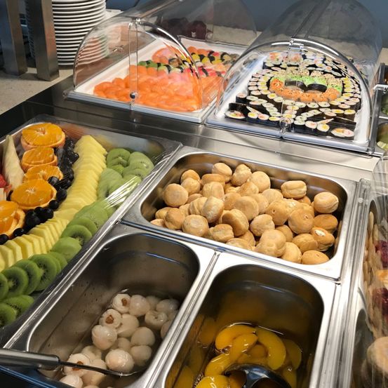 Ein Buffettisch hat verschiedene Essensschalen, darunter Sushi, Orangenscheiben, Kiwis und andere Früchte. Die Schalen haben transparente Deckel. Eine Schale enthält Brot.