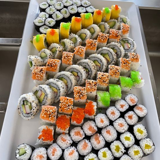 Ein Tablett mit Sushi mit verschiedenen Füllungen, einschließlich Gurke, Avocado und Lachs, garniert mit Sesam und Rogen. Die Sushi sind ordentlich in Reihen auf einem weißen Tablett angeordnet.