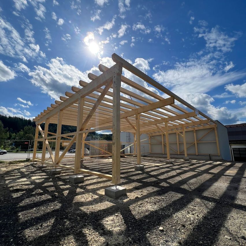 Ein Holzbau im Bau auf einem sonnigen Tag mit blauem Himmel und Wolken. Die Struktur wird von Holzbalken und Säulen gestützt.