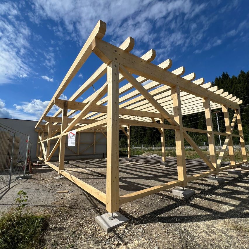 Eine Holzkonstruktion im Bau, gestützt von Holzbalken und Betonblöcken, mit einem Schild links und einem Gebäude im Hintergrund.