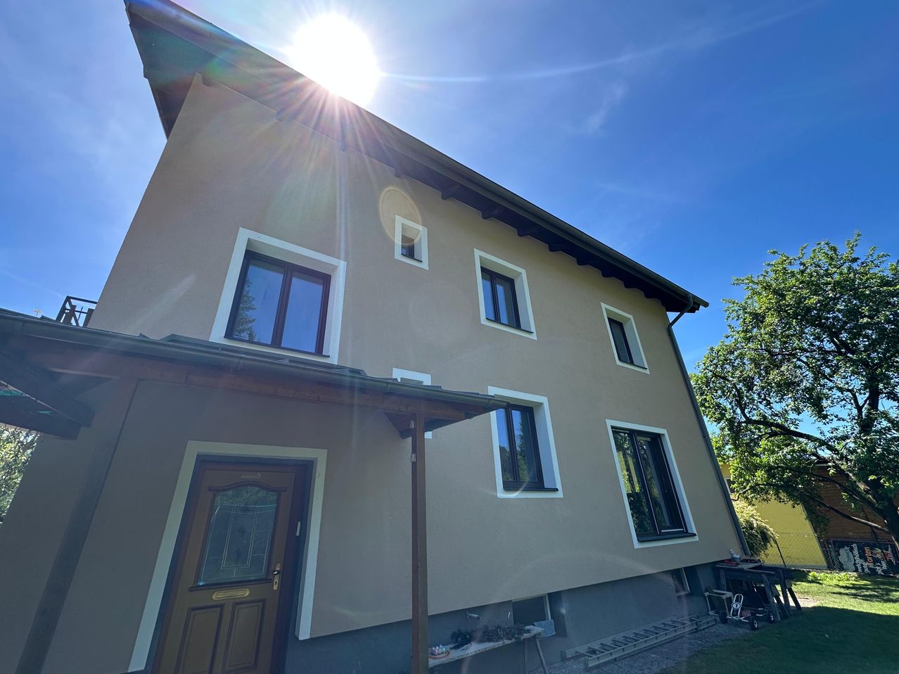 Ein großes Haus mit brauner Außenfassade, einem sonnenbeschienenen Dach und weiß umrandeten Fenstern. Die Tür ist braun mit einem Glasfenster. Ein Auto ist davor geparkt.