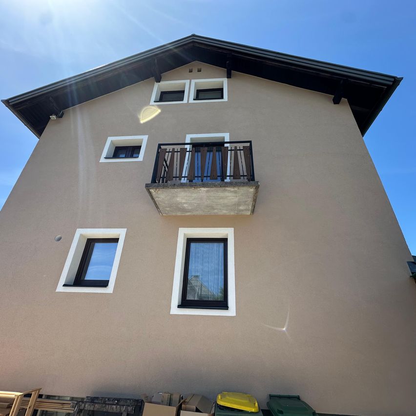 Ein dreistöckiges Gebäude mit beigefarbener Fassade, weißen Fensterumrandungen und einem Balkon im zweiten Stock. Unten befinden sich Kisten und Mülleimer.