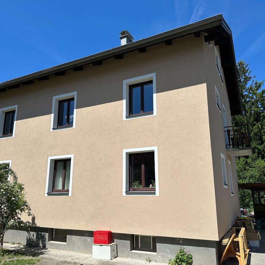 Ein beiges Haus mit weißen Fensterrahmen, mehreren Fenstern und einem Balkon. Ein roter Aufbewahrungsbehälter steht auf dem Boden unter einem der Fenster.