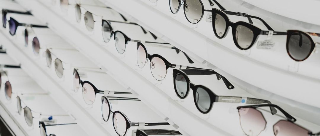 Eine Sammlung von Sonnenbrillen in verschiedenen Farben und Stilen, ordentlich auf einem weißen Regal angeordnet und ihre Gläser und Rahmen präsentierend.