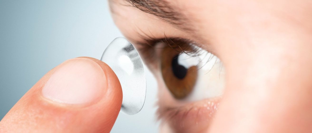 Eine Nahaufnahme eines Augens mit einem Finger, der eine transparente Kontaktlinse hält. Das Auge hat braune Iris und Wimpern.