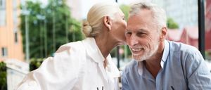 Ein älteres Paar sitzt am Tisch und hält Händchen. Die Frau küsst den Mann auf die Wange. Sie hält ein Glas Orangensaft mit Strohhalm.