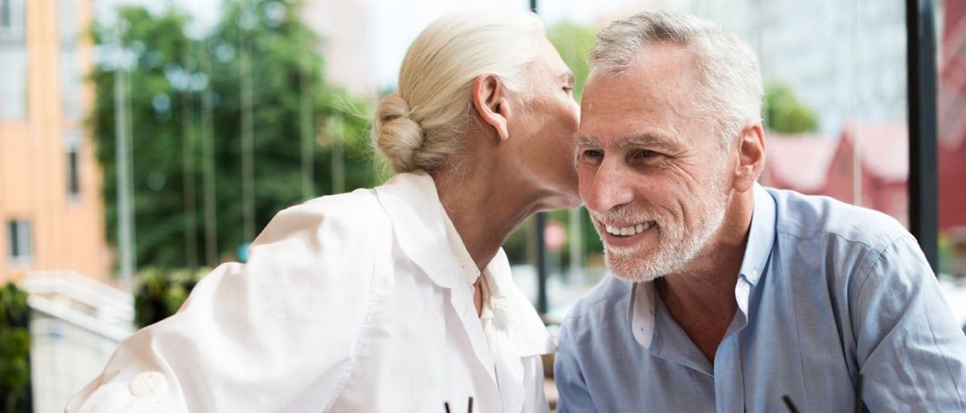 Ein älteres Paar sitzt am Tisch und hält Händchen. Die Frau küsst den Mann auf die Wange. Sie hält ein Glas Orangensaft mit Strohhalm.