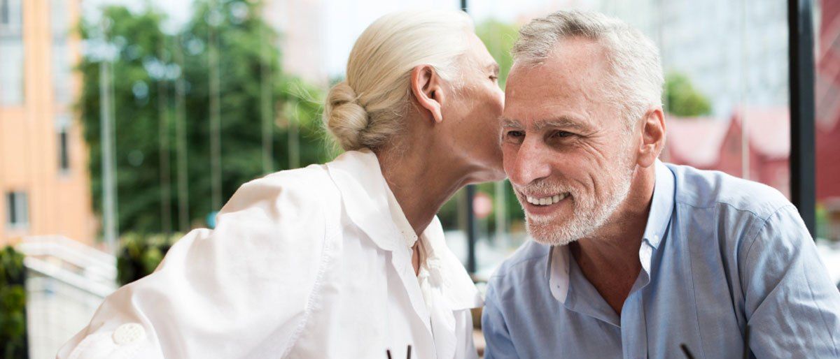 Ein älteres Paar sitzt am Tisch und hält Händchen. Die Frau küsst den Mann auf die Wange. Sie hält ein Glas Orangensaft mit Strohhalm.