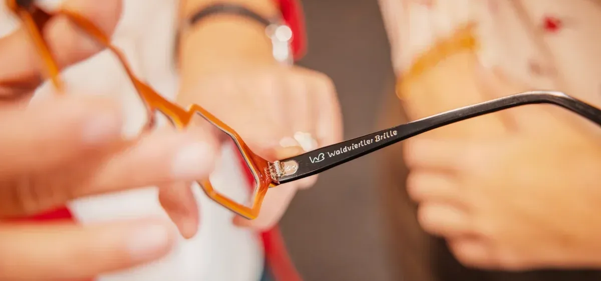 Ein Paar Brille mit orangefarbenen Gläsern wird von einer Person gehalten. Die Brille hat den Text 'Waldviertler Brille' auf der Seite.