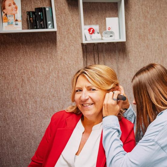 Eine Frau lässt sich die Ohren von einer anderen Frau piercen, lächelt und schaut sie an. Dahinter befinden sich Regale mit Büchern, Produkten und Kopfhörern.