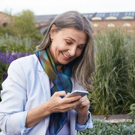 Eine ältere Frau mit grauen Haaren lächelt und schaut auf ihr Handy in einem Garten.